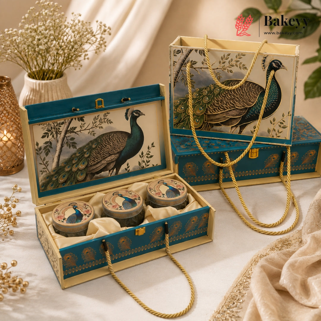 A decorative gift set displayed on a draped fabric surface, featuring an open teal and cream box with peacock artwork, three round tin containers inside, a matching tote bag with rope handles, and additional matching boxes in the background. The scene includes styled props such as dried flowers, a candle holder, and soft fabric drapery.