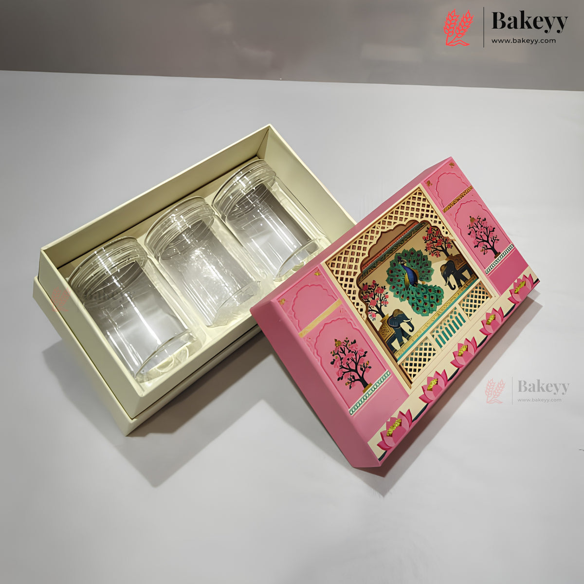 Three clear glass cups presented in a cream-colored tray inside a pink decorative gift box, with the illustrated lid placed beside it on a light gray surface.