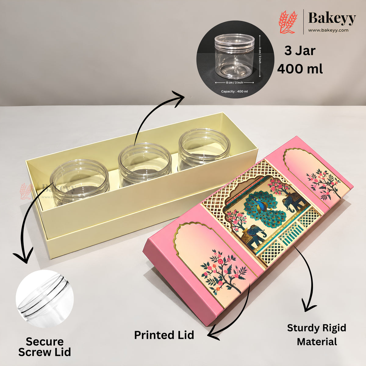 Three clear glass jars with screw-on lids displayed in a cream-colored presentation box, alongside a pink decorative printed lid and callout arrows explaining features. The Bakeyy brand logo and website are visible in the top right.