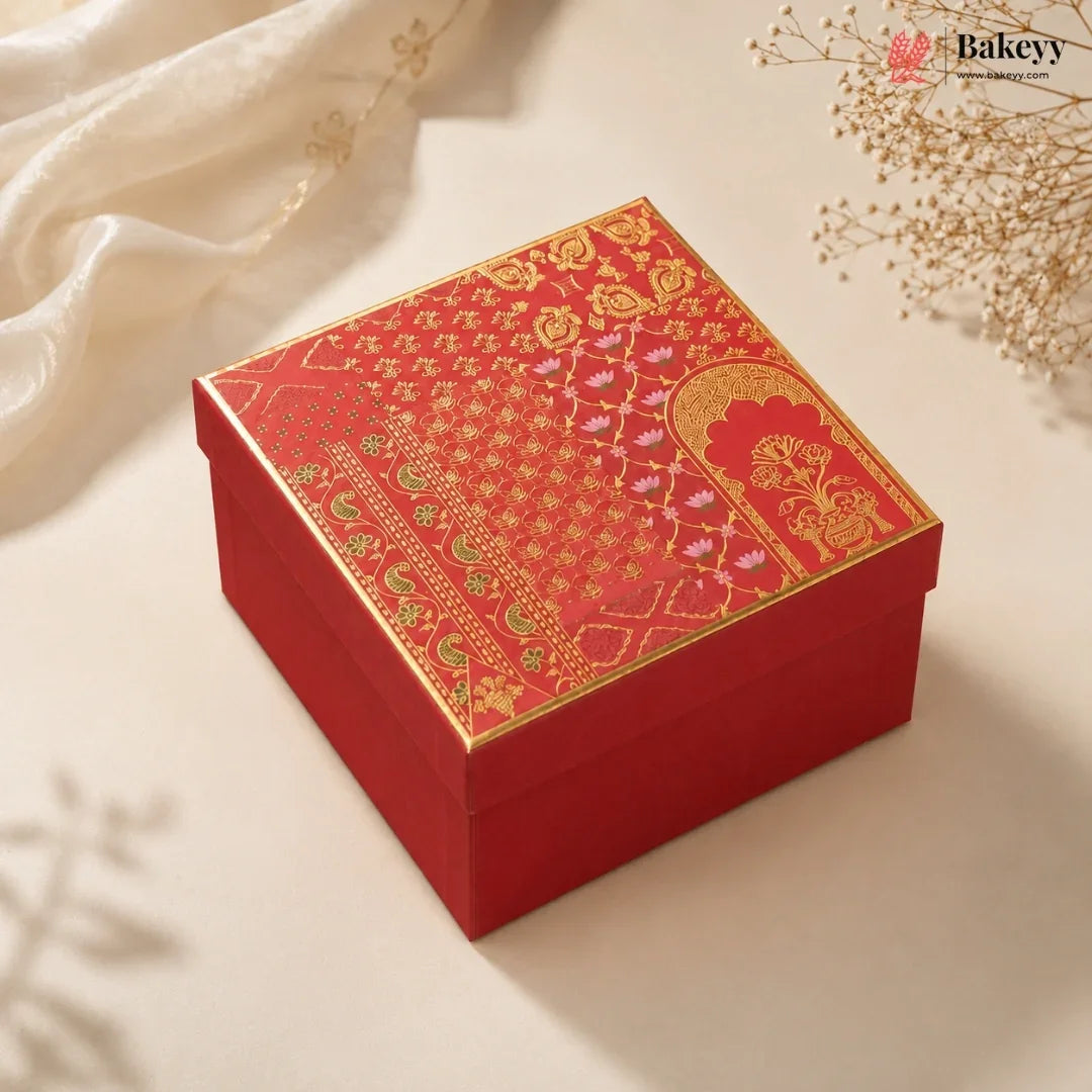 A square red gift box with an ornate gold and pink patterned lid, styled on a light beige surface with soft white fabric and dried floral decorations.