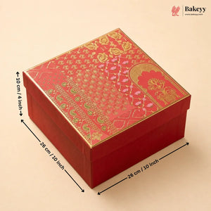 A red square gift box with ornate gold foil patterns on the lid, shown with black dimension arrows and text indicating measurements of 26 cm by 26 cm by 10 cm. The Bakeyy logo appears in the top right corner.