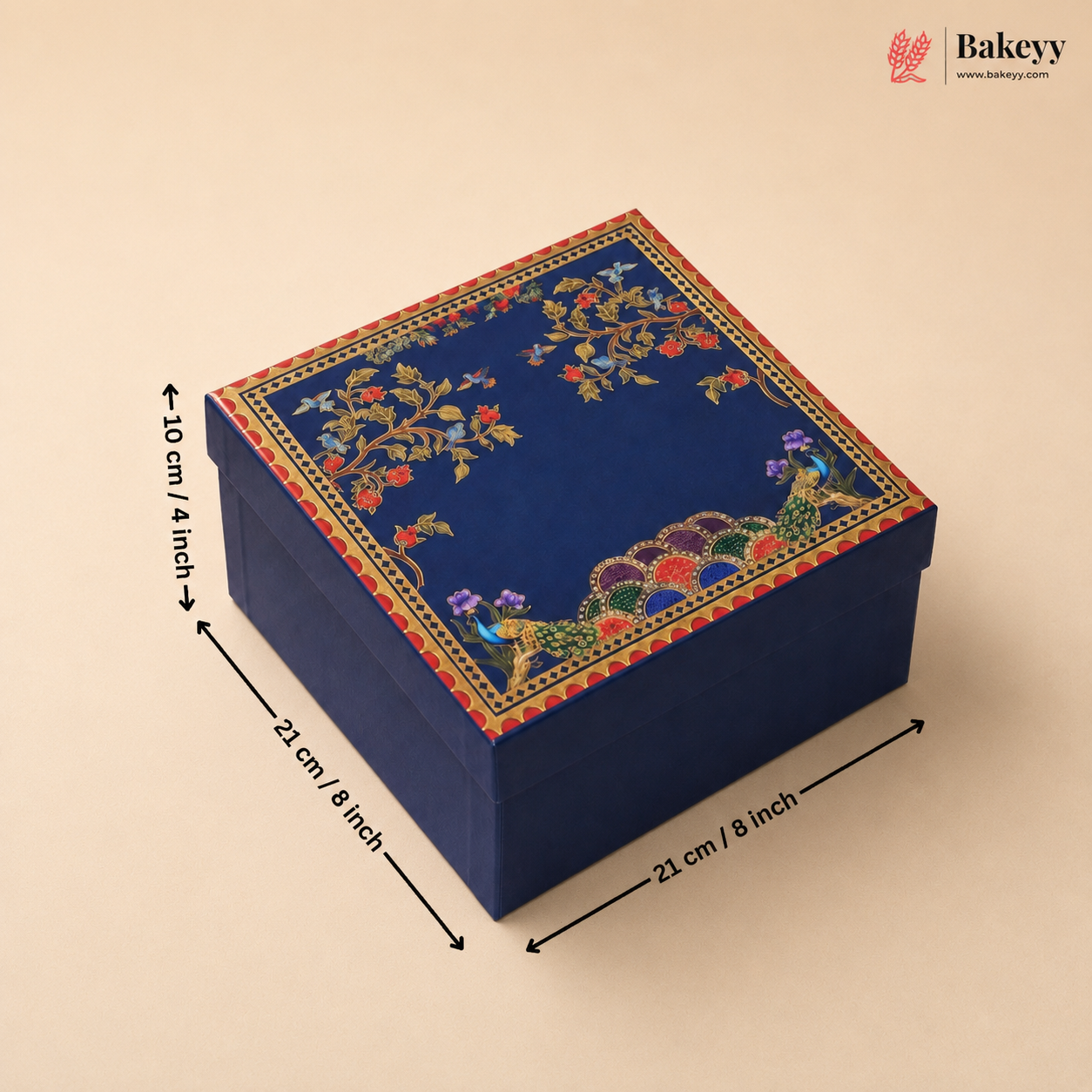 A square dark blue decorative gift box with floral and peacock embroidery on the lid, shown with dimension arrows and labels indicating 21 cm by 21 cm by 10 cm. The Bakeyy logo appears in the top right corner.