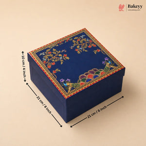 A square dark blue decorative gift box with floral and peacock embroidery on the lid, shown with dimension arrows and labels indicating 21 cm by 21 cm by 10 cm. The Bakeyy logo appears in the top right corner.