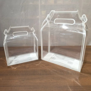 Two clear plastic gift boxes with integrated handles, shown in two different sizes on a wooden surface against a neutral background.