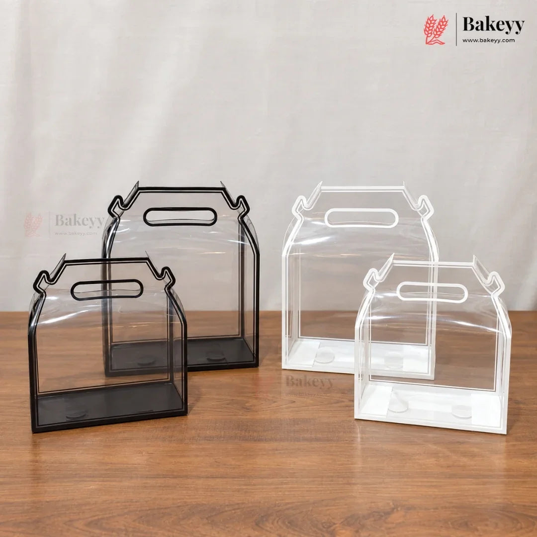 Three clear plastic bakery gift boxes with integrated handles, shown in black and white frames on a wooden table against a neutral fabric backdrop. The Bakeyy logo is visible in the top right corner.