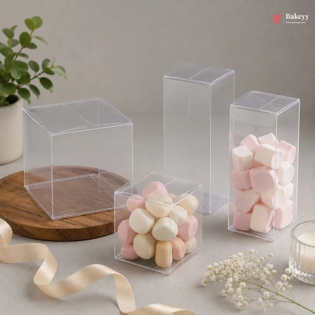 Clear plastic boxes containing pink and white macarons on a neutral background with a plant and ribbon.