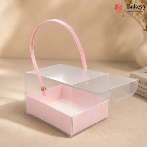 Clear gift box with pink handle and interior on a beige surface, featuring the Bakeyy brand.