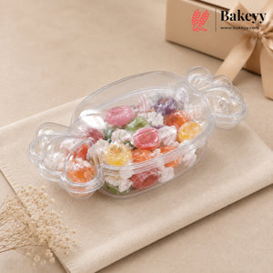 A clear plastic candy dish filled with assorted colorful candies, styled on a beige fabric napkin with dried flowers and a gift box in the background. The Bakeyy brand logo is visible in the top right corner.