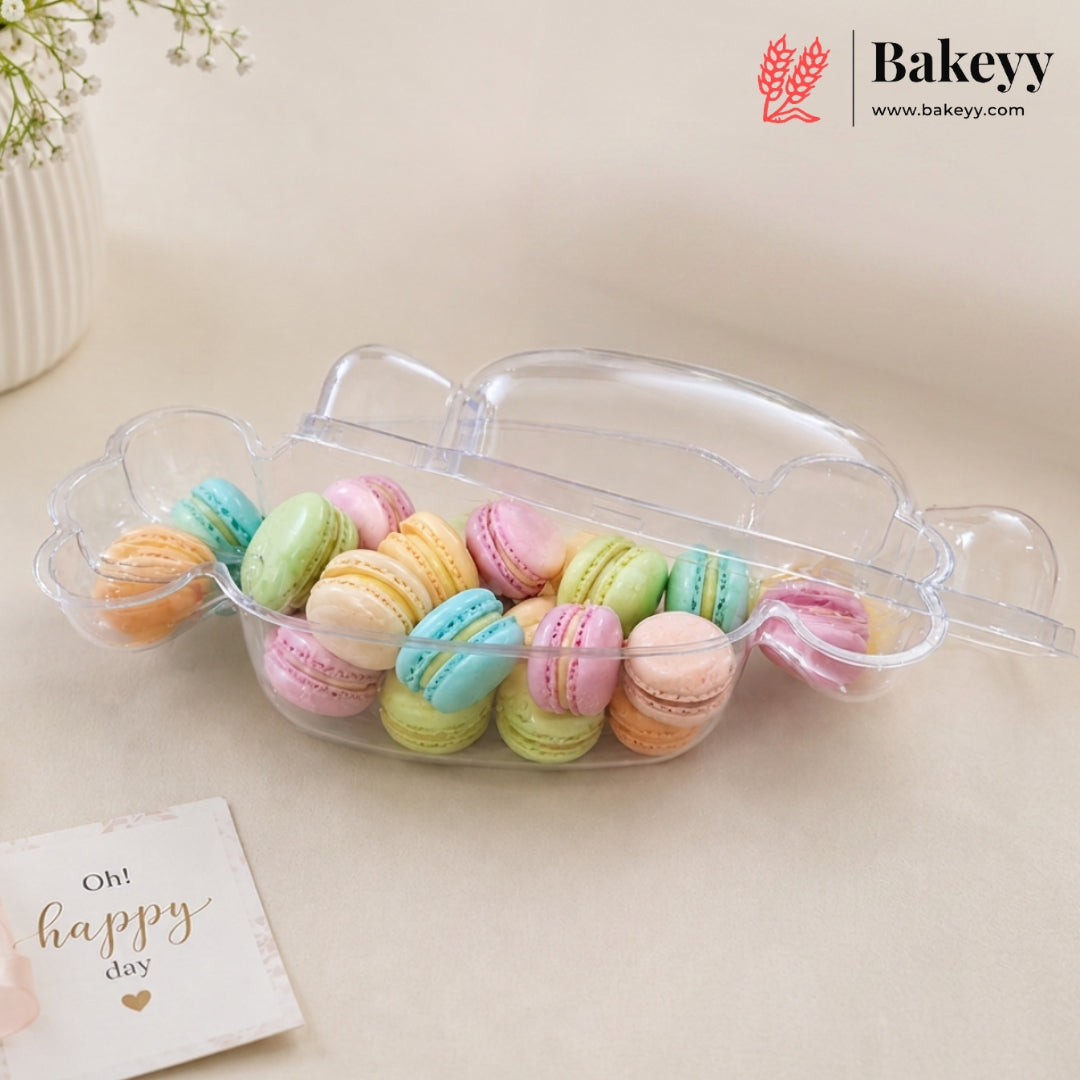 A clear plastic macaron container with a lid, filled with colorful pastel macarons, on a beige surface with a small card that reads 'Oh! happy day' and a Bakeyy logo in the top right corner.