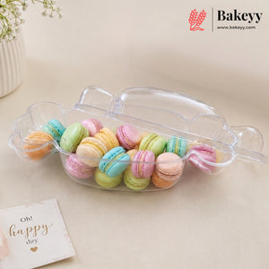 A clear plastic macaron container with a lid, filled with colorful pastel macarons, on a beige surface with a small card that reads 'Oh! happy day' and a Bakeyy logo in the top right corner.