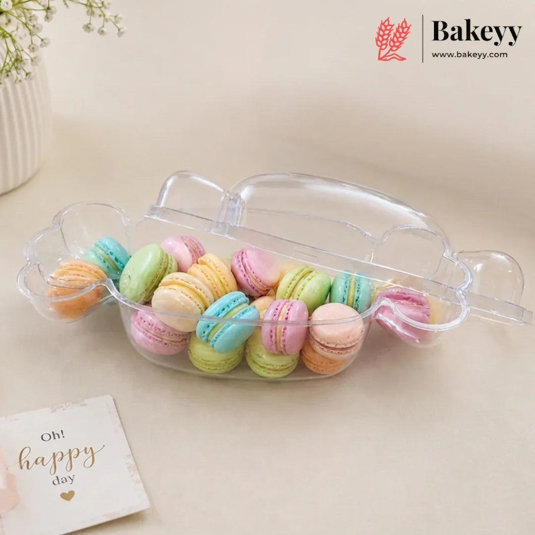 A clear plastic macaron container with a lid, filled with colorful pastel macarons, on a beige surface with a small card that reads 'Oh! happy day' and a Bakeyy logo in the top right corner.