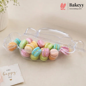 A clear plastic macaron container with a lid, filled with colorful pastel macarons, on a beige surface with a small card that reads 'Oh! happy day' and a Bakeyy logo in the top right corner.