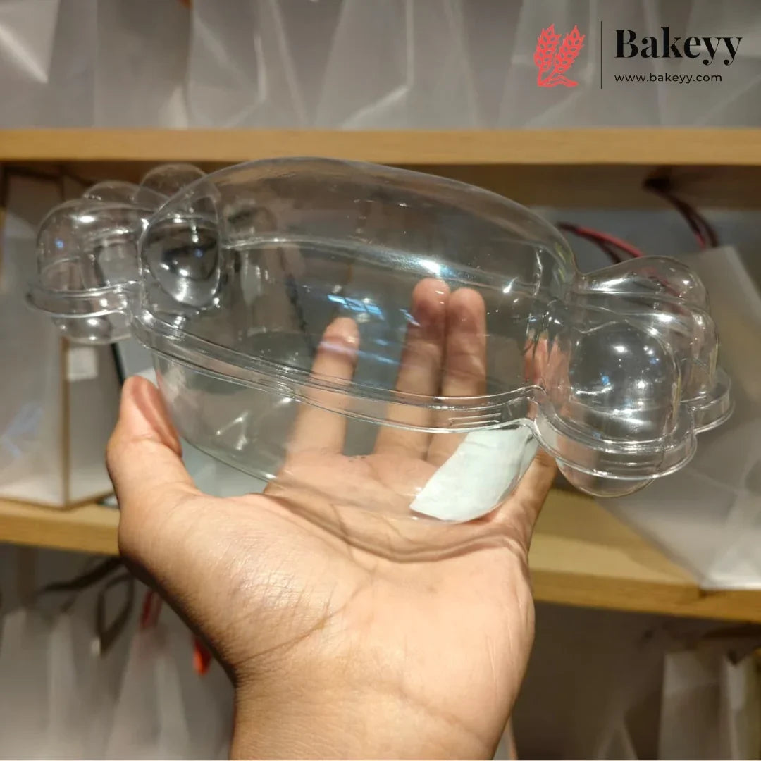 A hand holding a clear plastic bakery mold or tray with two small raised sections on the sides, shown against a shelf background with a Bakeyy logo in the top right.