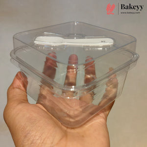 A hand holding a clear plastic square food container with a matching lid and a white plastic spoon resting inside, against a plain light background. The Bakeey logo is visible in the top right corner.
