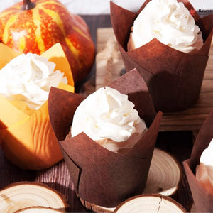Cupcakes with white frosting in brown paper cones on a wooden surface with pumpkins in the background.