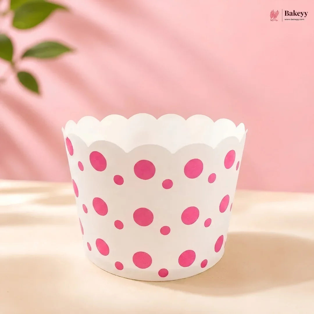 A white paper cupcake liner with pink polka dots, displayed on a light beige surface against a soft pink background with a leafy green accent in the upper left.