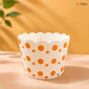 A white paper cupcake liner with orange polka dots, displayed on a beige surface with a warm yellow background and soft leaf shadows.