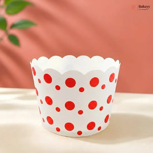 A white paper cupcake liner with red polka dots, standing on a light beige surface against a warm peach-pink background with soft leaf shadows.