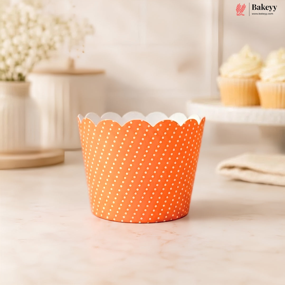 An orange paper cupcake liner with white polka dots and a scalloped edge, shown on a light marble countertop with a softly blurred kitchen background featuring cupcakes.