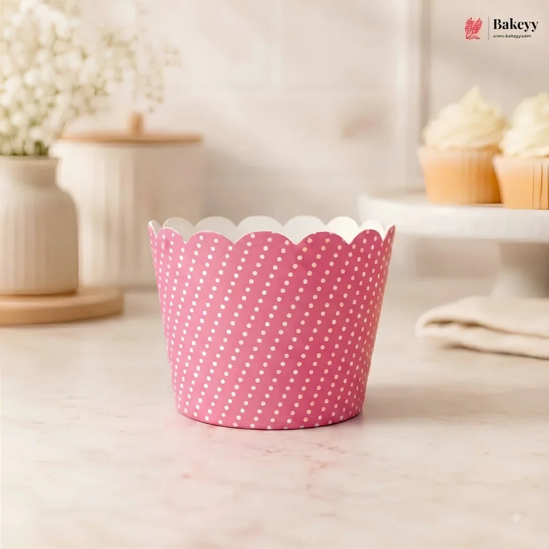 A pink paper cupcake liner with white polka dots and a scalloped edge, shown on a light marble countertop with a blurred bakery background featuring cupcakes and a Bakeyy logo in the top right.