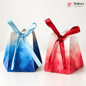 Two small paper favor boxes with watercolor-style gradients, one in blue and one in red, each tied with a satin ribbon bow, on a clean white background. The Bakeyy logo appears in the top right corner.