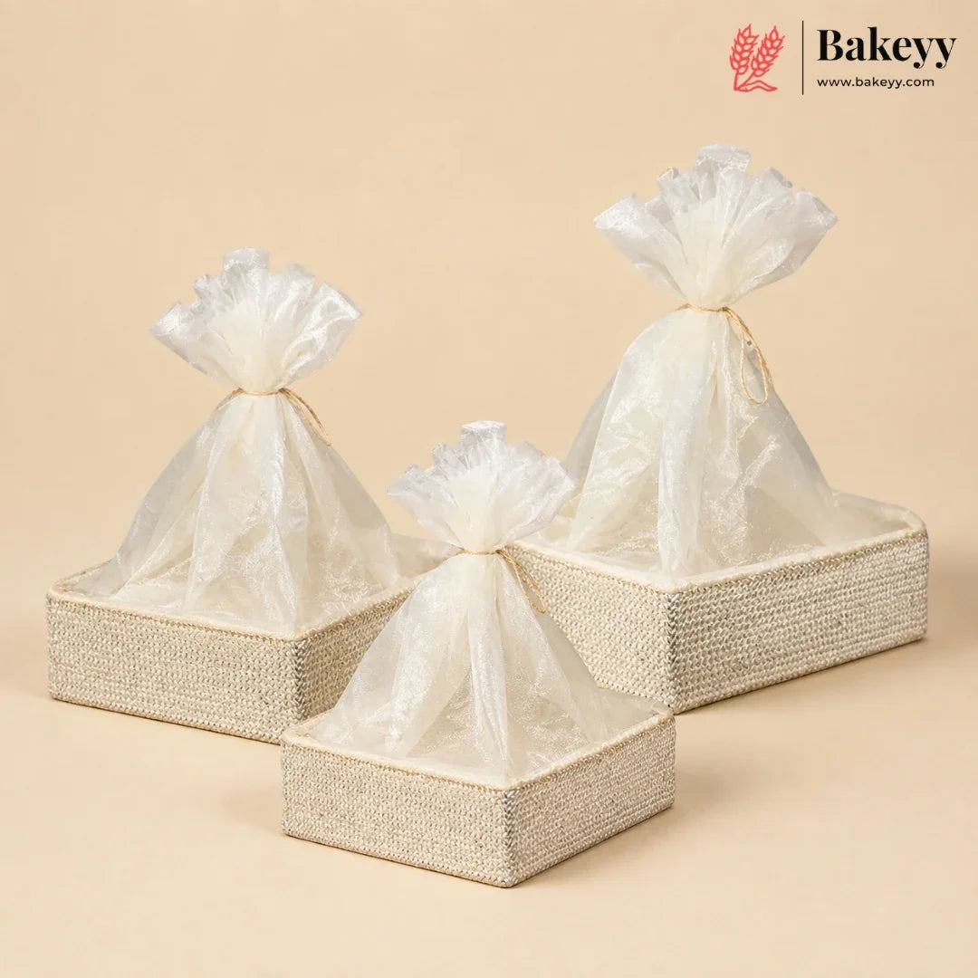 Three woven rectangular gift boxes in beige and cream, each holding a translucent cellophane drawstring bag tied at the top, arranged on a warm beige background. A Bakeyy logo appears in the upper right corner.