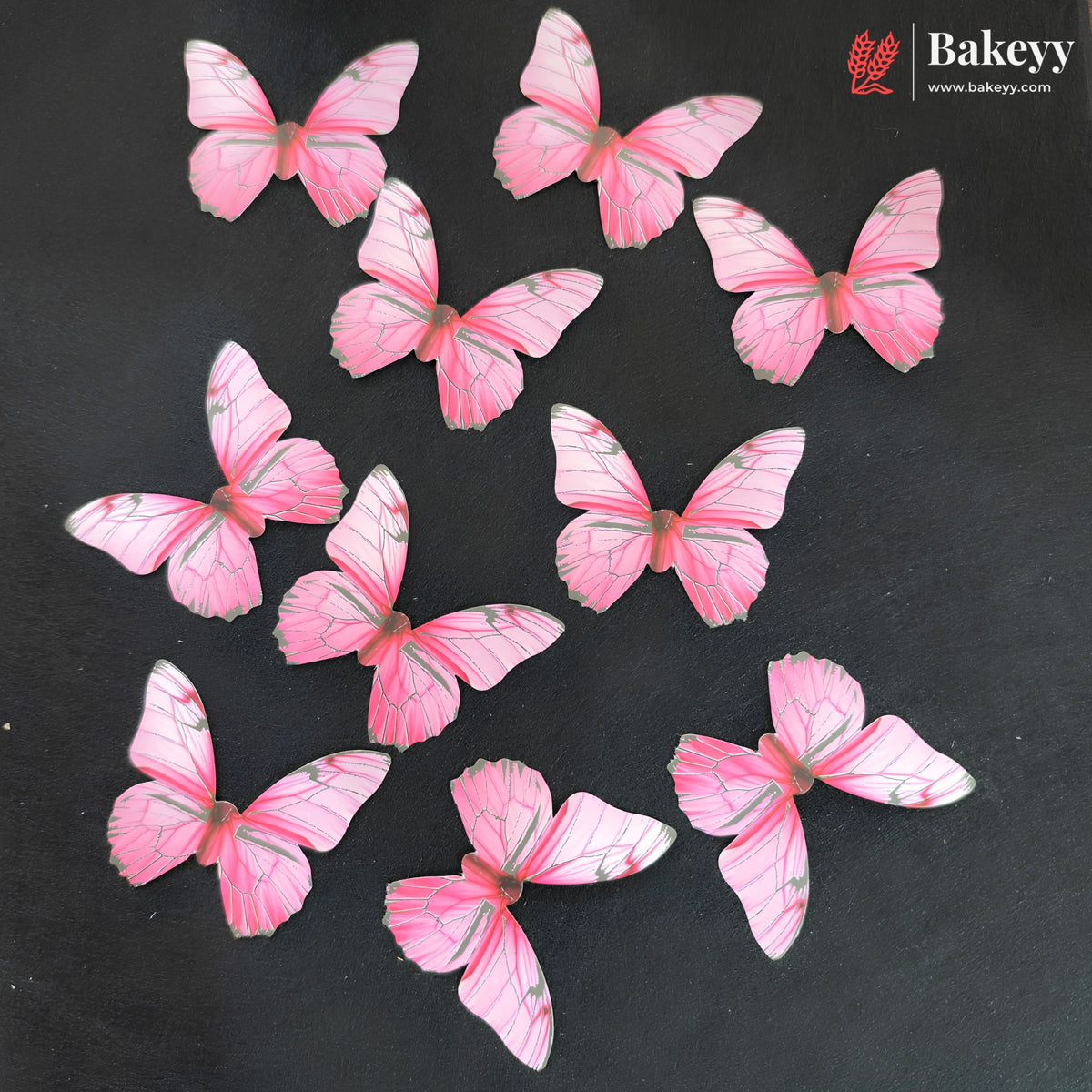 Nine pink butterfly-shaped cake toppers with gray vein details scattered on a dark gray background, with the Bakeyy logo in the top right corner.