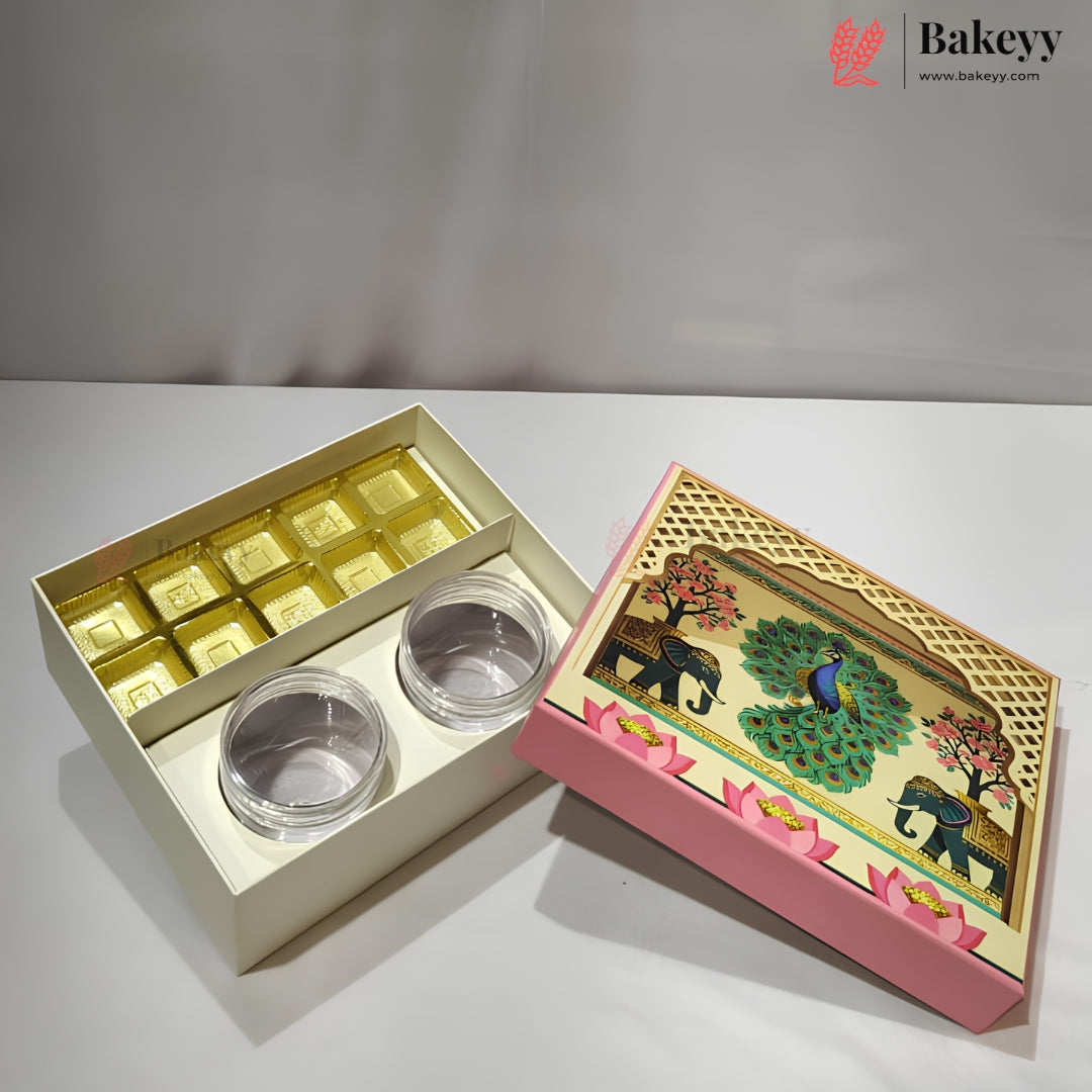 A decorative gift box set from Bakeyy, featuring an open cream-colored box with gold square-shaped items and two small clear round containers, alongside a pink and gold illustrated lid with elephants, peacocks, and lotus flowers.