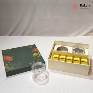 A bakery gift set featuring a dark green lotus-themed box lid, an open cream-colored box containing gold foil-wrapped treats and two clear round jars, and one loose clear jar on a white surface.