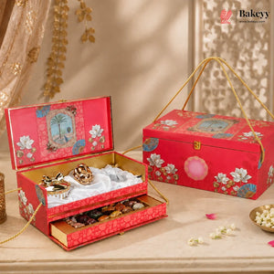 An ornate red gift box set for chocolates, shown open with gold-trimmed compartments holding assorted chocolates and sweets, alongside the closed matching box on a beige surface with decorative floral props.