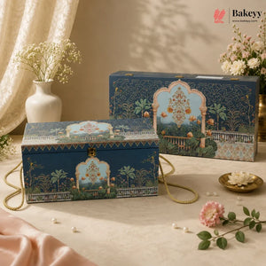 Two decorative blue bakery gift boxes with gold handles and ornate illustrated arch motifs, styled on a beige fabric surface with flowers and draped satin in a warm indoor setting. A Bakeyy watermark/logo appears in the top right.