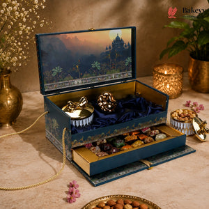 An ornate blue and gold gift box set displayed on a beige tablecloth, featuring a decorative illustrated lid, plush velvet compartments, and an open drawer filled with assorted chocolates and nuts. The scene is styled with candles, flowers, and a plant, and the Bakevy logo appears in the top right corner.
