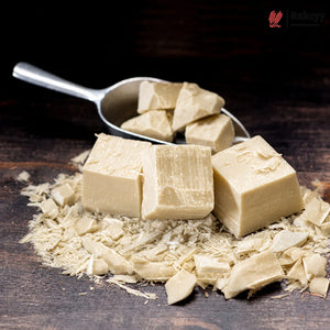 Four pale beige candy or soap blocks with shavings scattered around them and more pieces in a metal scoop, photographed on a dark wooden surface. A small Bakeery watermark appears in the top-right corner.