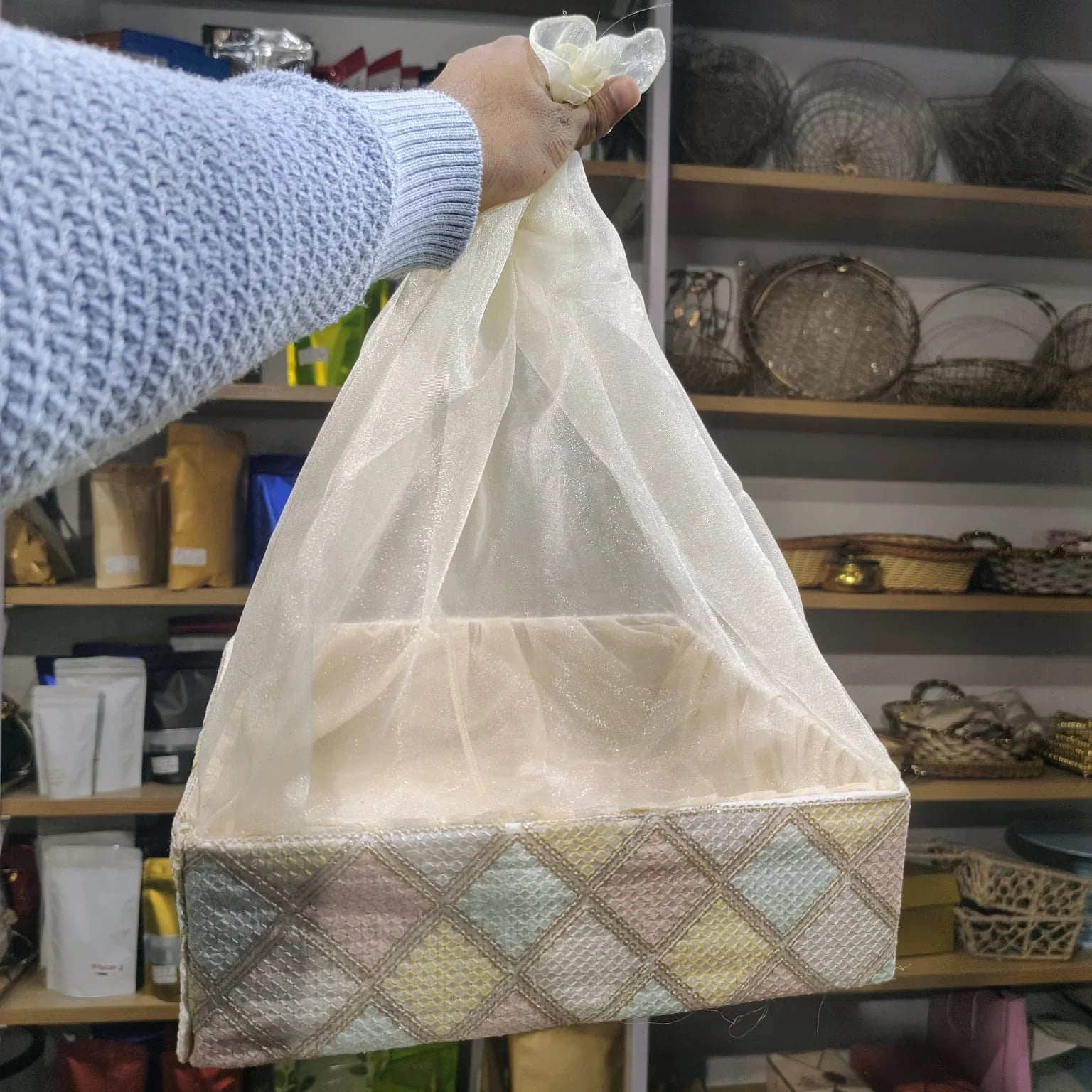 Hand holding a patterned box with a sheer white cover against a background of shelves with various items.