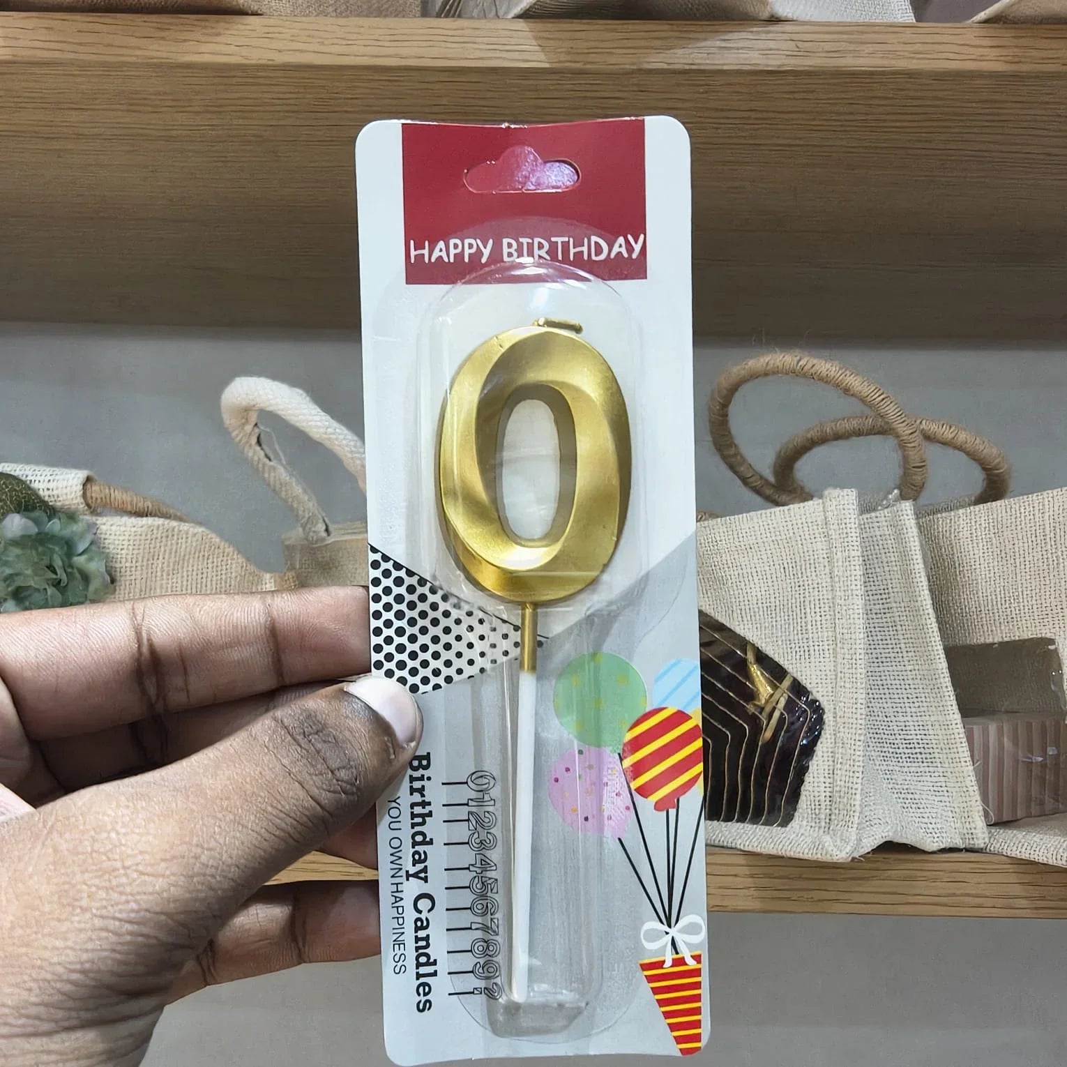A hand holds a gold metal letter 'O' birthday candle in its retail packaging, which features a red 'HAPPY BIRTHDAY' header and colorful balloon graphics.