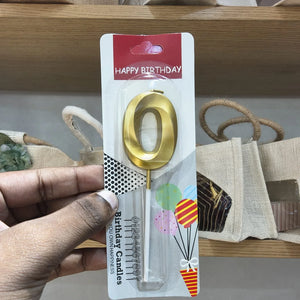 A hand holds a gold metal letter 'O' birthday candle in its retail packaging, which features a red 'HAPPY BIRTHDAY' header and colorful balloon graphics.