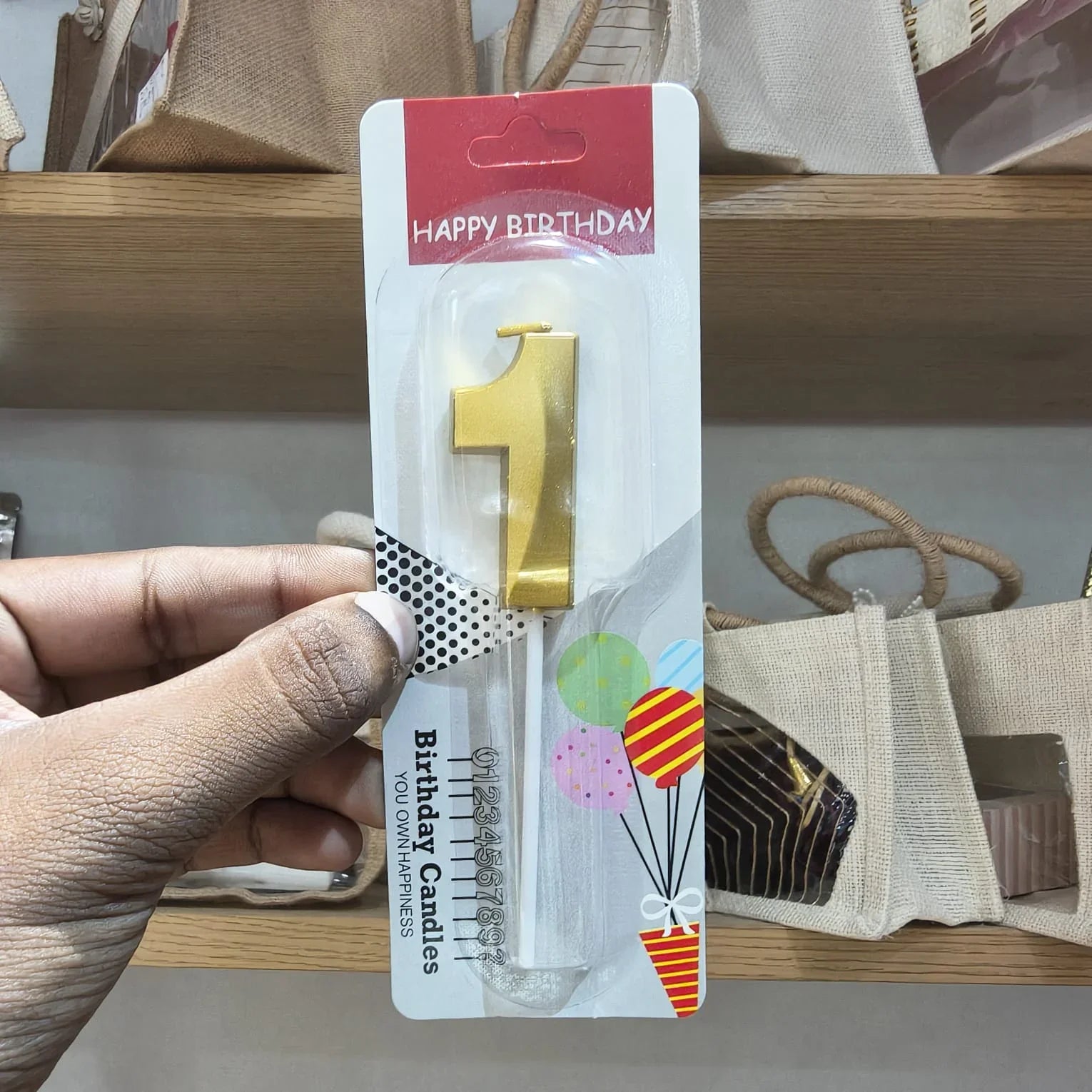 A hand holding a packaged gold number 1 birthday candle in a clear plastic holder with a red 'HAPPY BIRTHDAY' header card and white backing, photographed on a store shelf.