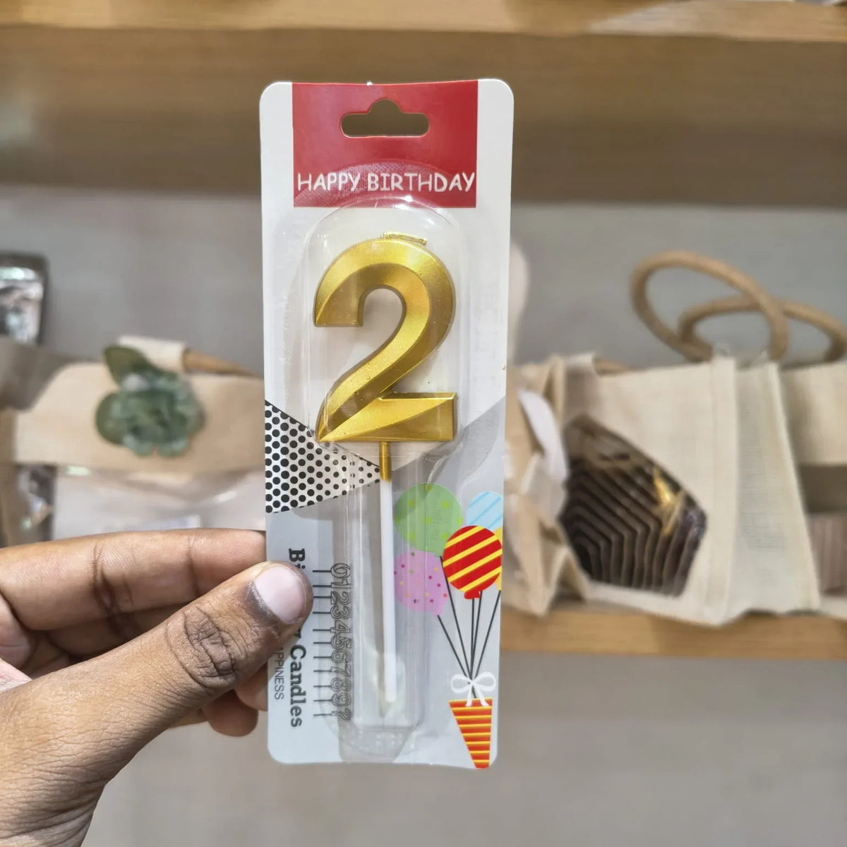 A hand holding a packaged gold plastic birthday candle shaped like the number 2, with 'HAPPY BIRTHDAY' printed on the card packaging.