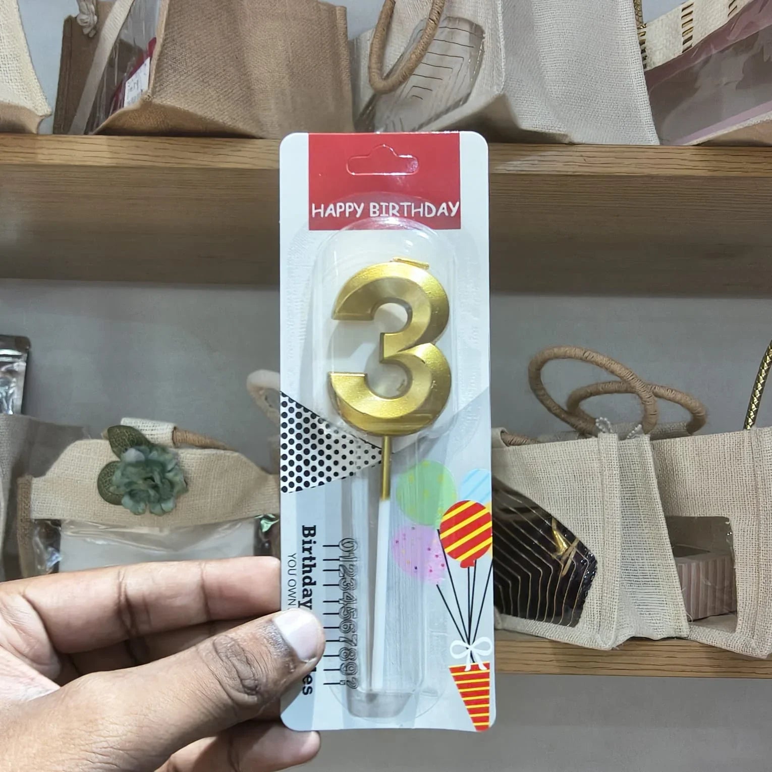 A hand holding a packaged gold number 3 birthday candle with a red 'HAPPY BIRTHDAY' header card, shown in a store setting with beige tote bags on wooden shelves in the background.