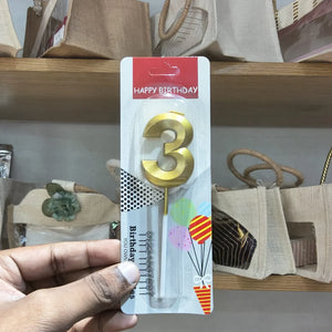 A hand holding a packaged gold number 3 birthday candle with a red 'HAPPY BIRTHDAY' header card, shown in a store setting with beige tote bags on wooden shelves in the background.