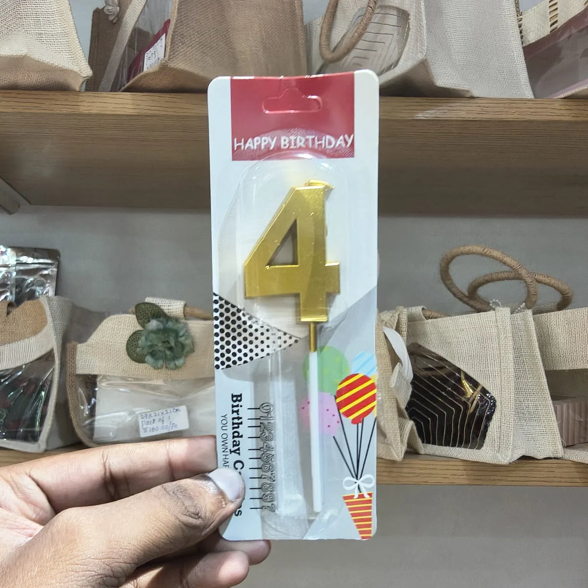 A hand holding a packaged gold birthday candle shaped like the number 4, with a red 'Happy Birthday' header and birthday-themed graphics on the card.