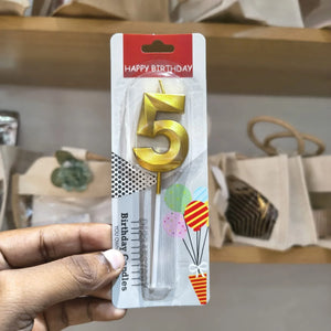 A hand holding a packaged gold number 5 birthday candle with a clear plastic base on a retail display shelf.