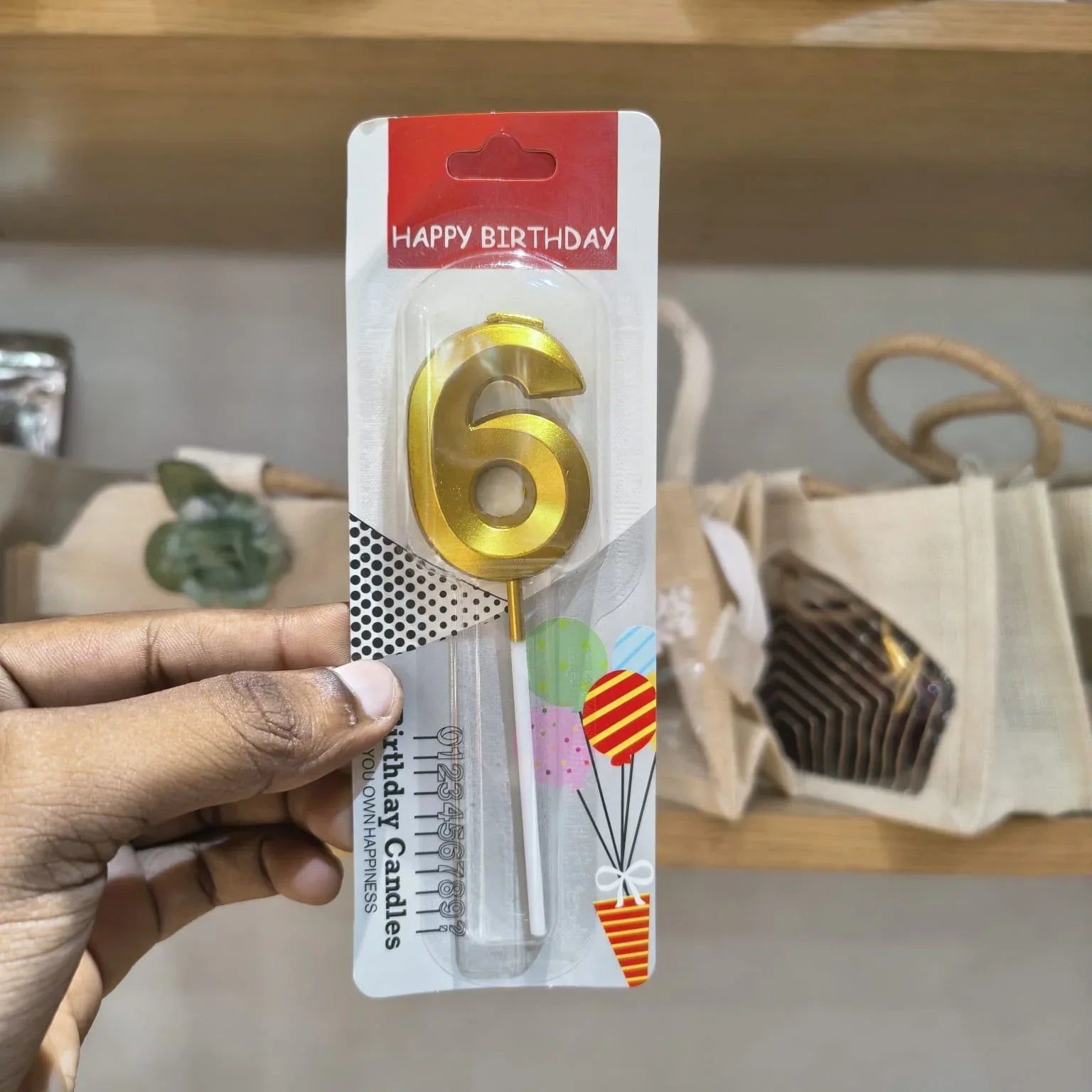 A hand holding a packaged gold number 6 birthday candle on a white backing card with a red 'HAPPY BIRTHDAY' header, against a blurred indoor background with shelves and bags.