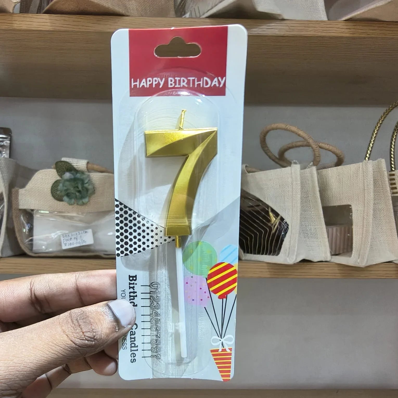 A hand holds a gold number 7 birthday candle in its original retail packaging, which features a red 'HAPPY BIRTHDAY' header and birthday-themed graphics, against a blurred background of shelves and bags.