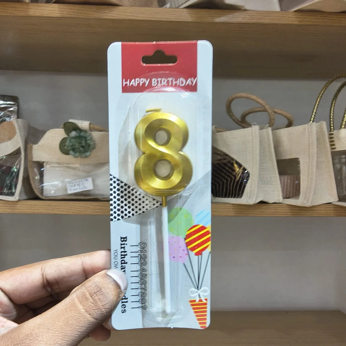A hand holding a packaged gold number 8 birthday candle on a retail display card, with shelves and bags in the background.