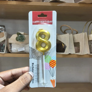 A hand holding a packaged gold number 8 birthday candle on a retail display card, with shelves and bags in the background.