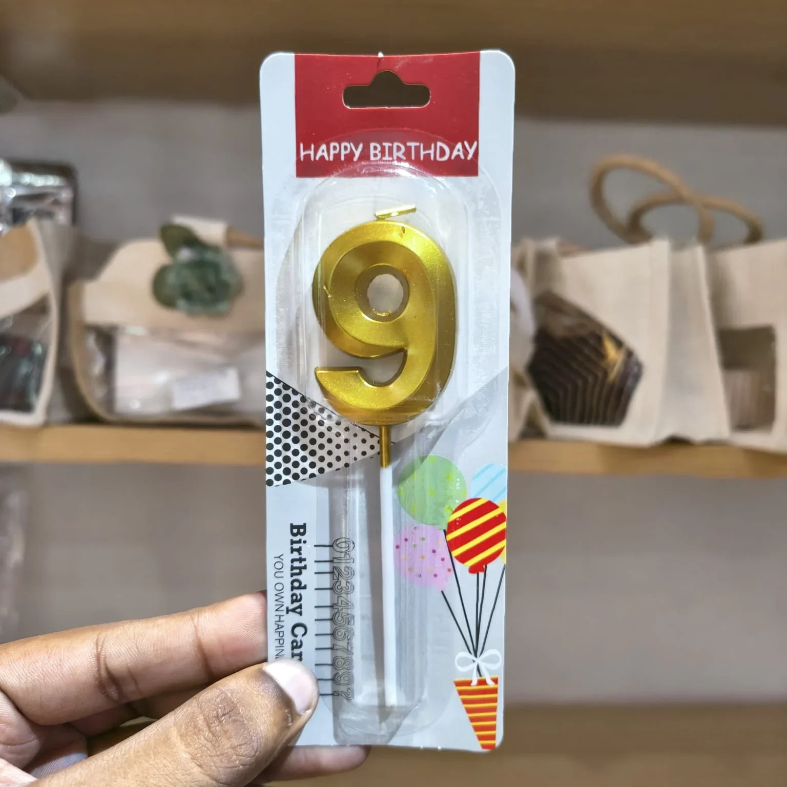 A hand holding a blister pack containing a gold metal number 9 birthday candle, with a red 'HAPPY BIRTHDAY' header and colorful balloon graphics on the card backing.