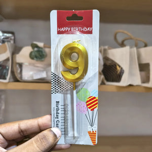 A hand holding a blister pack containing a gold metal number 9 birthday candle, with a red 'HAPPY BIRTHDAY' header and colorful balloon graphics on the card backing.