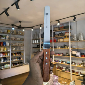 A hand holding a serrated bread knife with a silver metal blade and a brown wooden handle, set against a blurred background of a retail store interior.