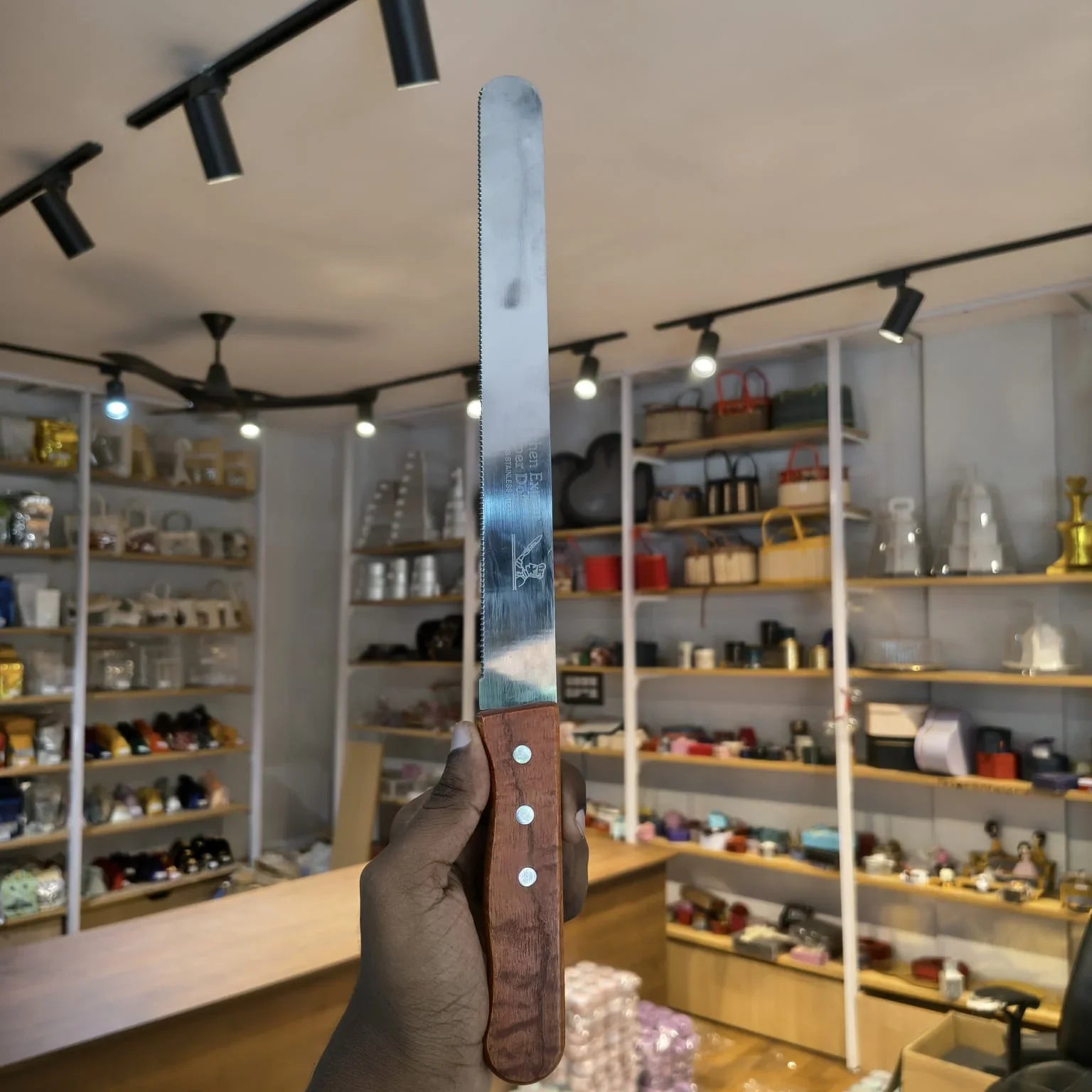 A hand holding a long serrated knife with a silver metal blade and a brown wooden handle, with a blurred retail store interior in the background.